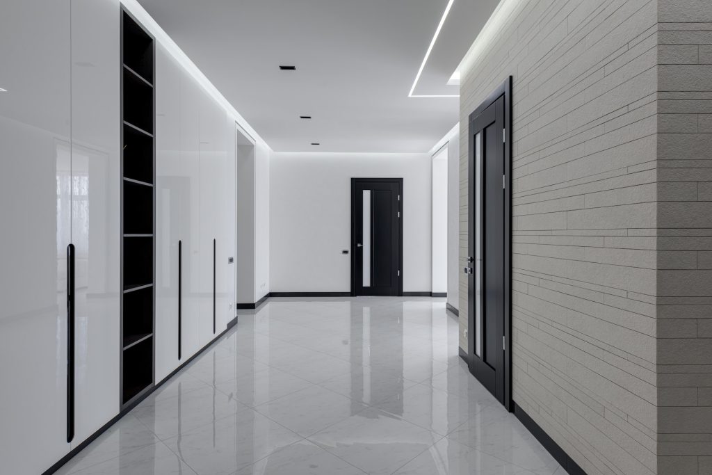 Wide corridor in modern house with white tiled walls and floor black wooden doors and shelves for storage built in wall